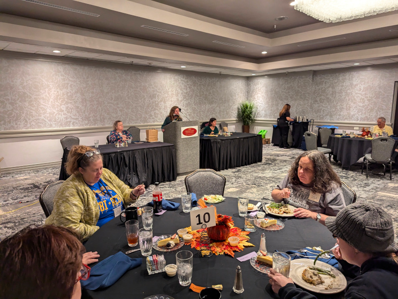 Kari Goodman seated at front round table with decorative Fall centerpiece in the middle of the table. Behind Kari is the front of the Ballroom where Kolby Garrison is standing behind the podium and talking into the microphone. Rita Kersh, President, ACBI (left) and Katie Frederick, President, ACBO (right) are seated at the tables.